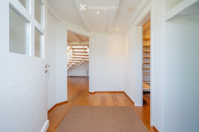 Bright, modern hallway with wooden flooring, white walls, and recessed lighting. Features a built-in shelving unit on the right and a staircase leading upwards in the background.