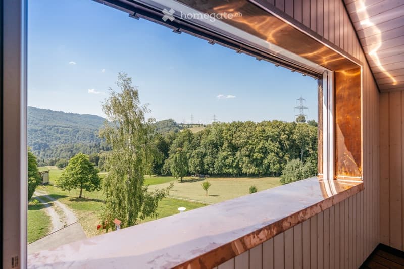 Balcony with wooden railings and a scenic view of a green valley, trees, and distant hills under a clear blue sky.