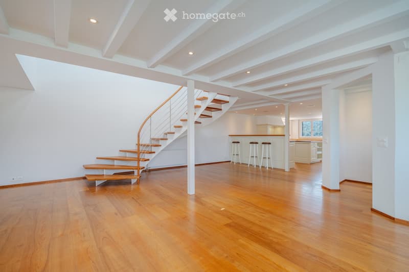 Open, airy space with polished hardwood floors, white walls, and a curved staircase leading upwards. The area includes a small seating area with bar stools and a kitchen counter in the background.