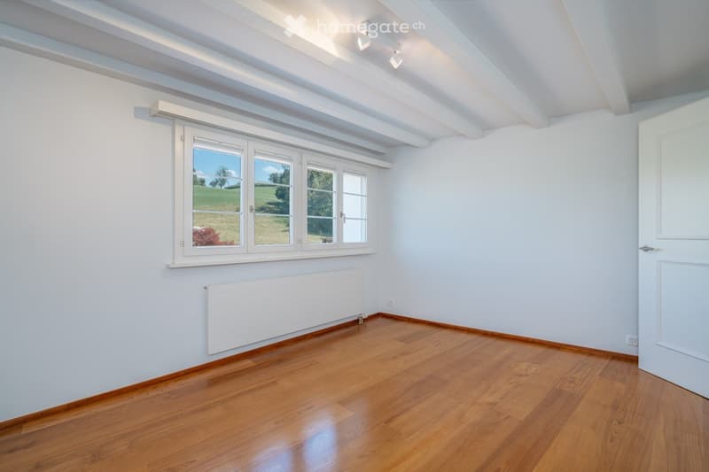 Empty room with wooden flooring, white walls, large window with white frame, radiator, and white door.