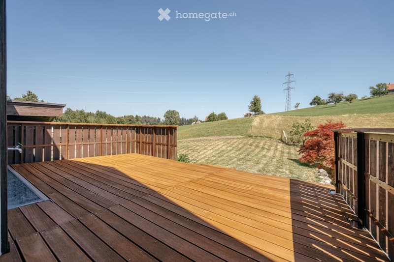 Wooden deck with railing, overlooking a grassy field and trees under a clear blue sky.