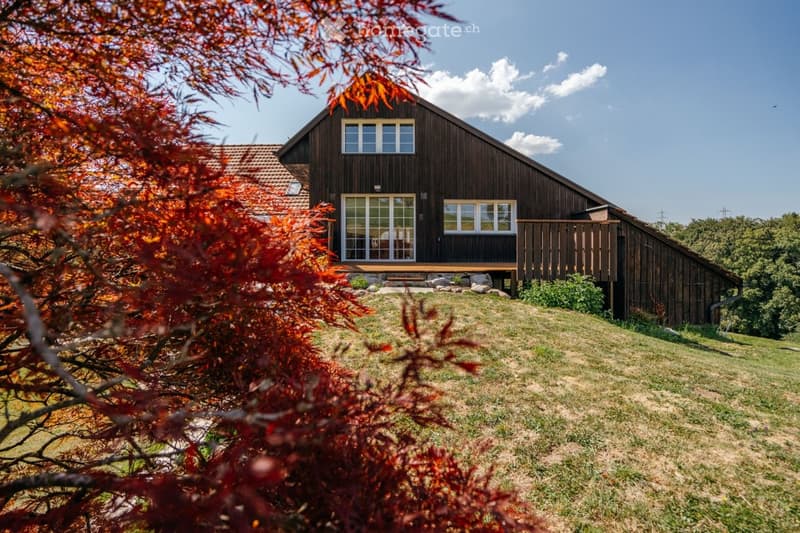 A two-story wooden house with a pitched roof, large windows, and a raised ground floor, surrounded by a grassy lawn and trees.