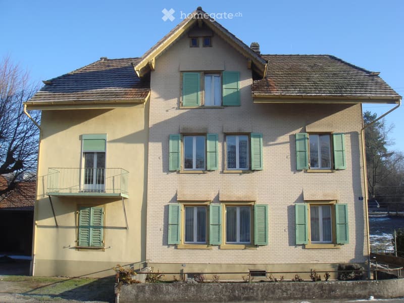 Two-story house with beige brick facade, green shutters, multiple windows, and a small balcony on the first floor.