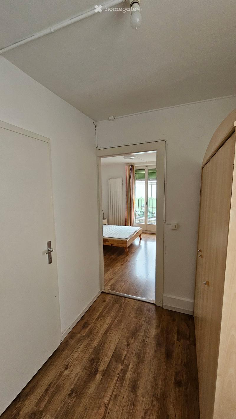 A narrow hallway with wooden flooring and white walls, leading to a bedroom with a bed and window view.