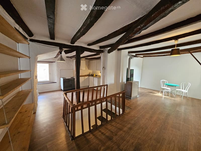 Open living space with exposed wooden beams, modern staircase, white walls, wooden floors, kitchen with white cabinets, central heating, window