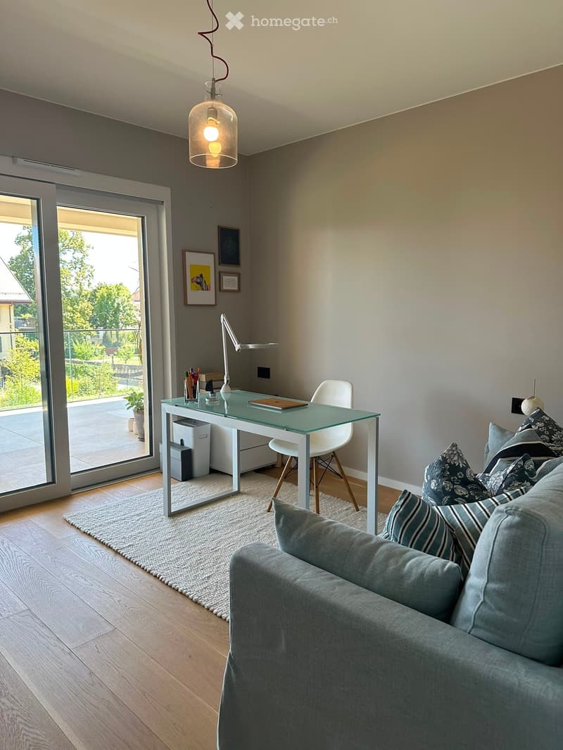 Modern living room with a grey sofa, coffee table, desk, and large windows leading to a balcony.