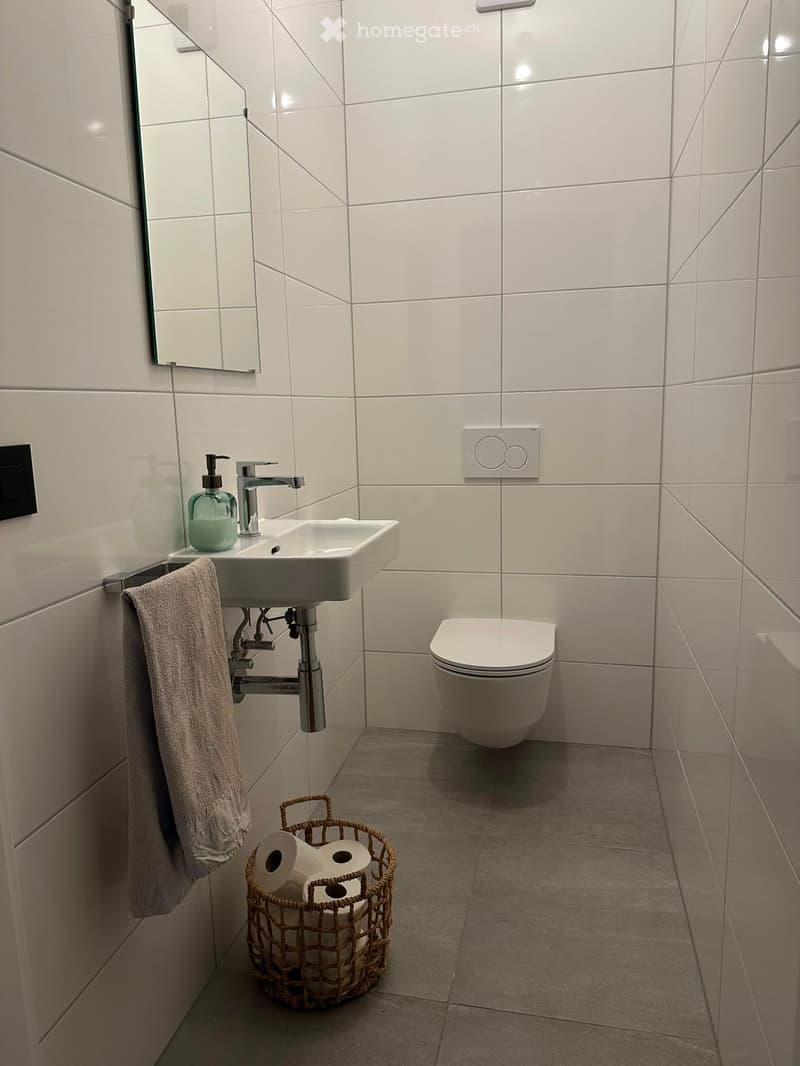 Modern bathroom with white tiles, a sink, a toilet, a towel, a mirror, and a basket with toilet paper.