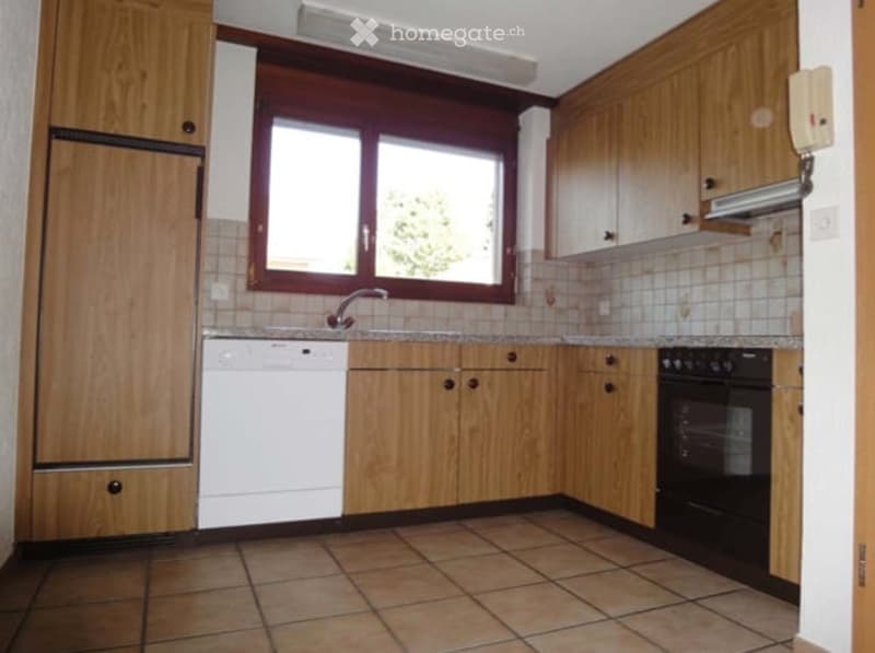 A well-lit kitchen with wooden cabinets, a tiled floor, a dishwasher, an oven, and a window above the sink.