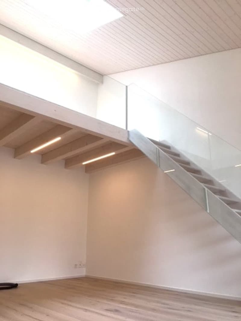 Empty room with wooden flooring, white walls, and a glass staircase leading to an upper level. The ceiling features recessed lighting and exposed wooden beams.