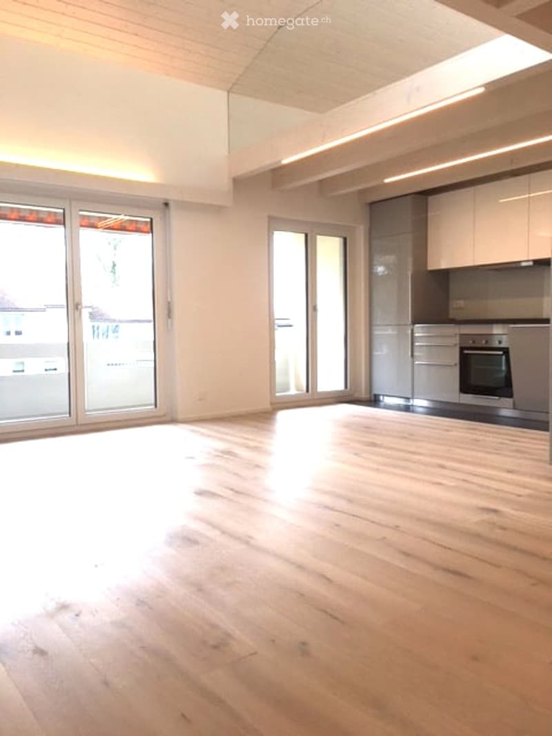 Bright, unfurnished room with wooden flooring, large glass doors leading to a balcony, and a small kitchen area with cabinets and appliances visible in the background.