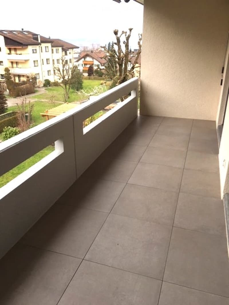 Balcony with tiled floor, white railing, and view of a green area with trees and neighboring buildings.