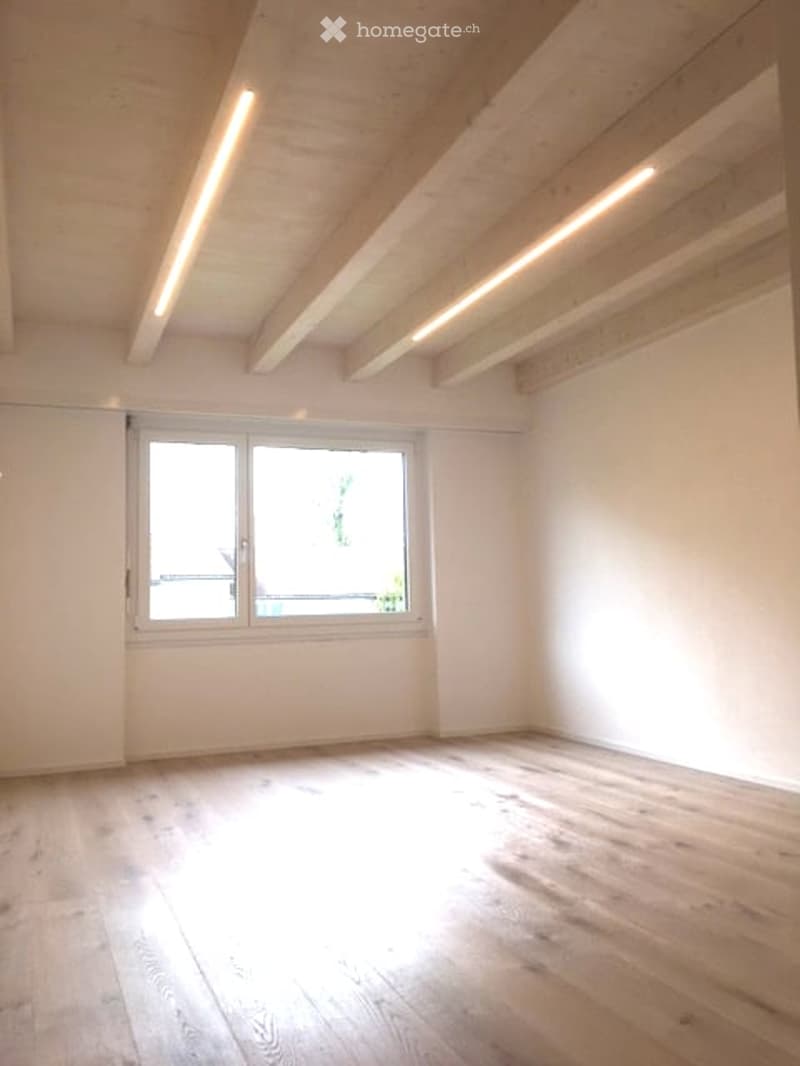 Completely empty room with white walls, light wooden floor, and large window letting in natural light. No furniture or equipment.