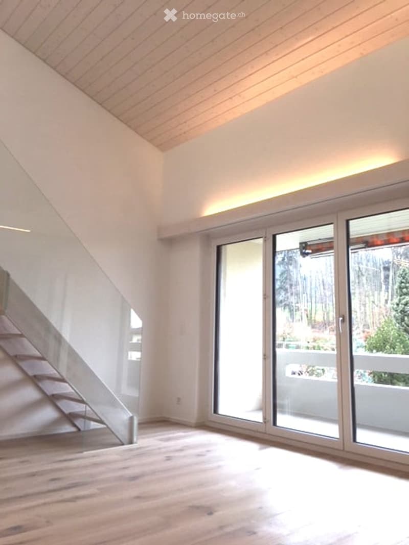 Bright, open room with wooden flooring, white walls, and large glass doors leading to a balcony. Minimalist design with recessed lighting.