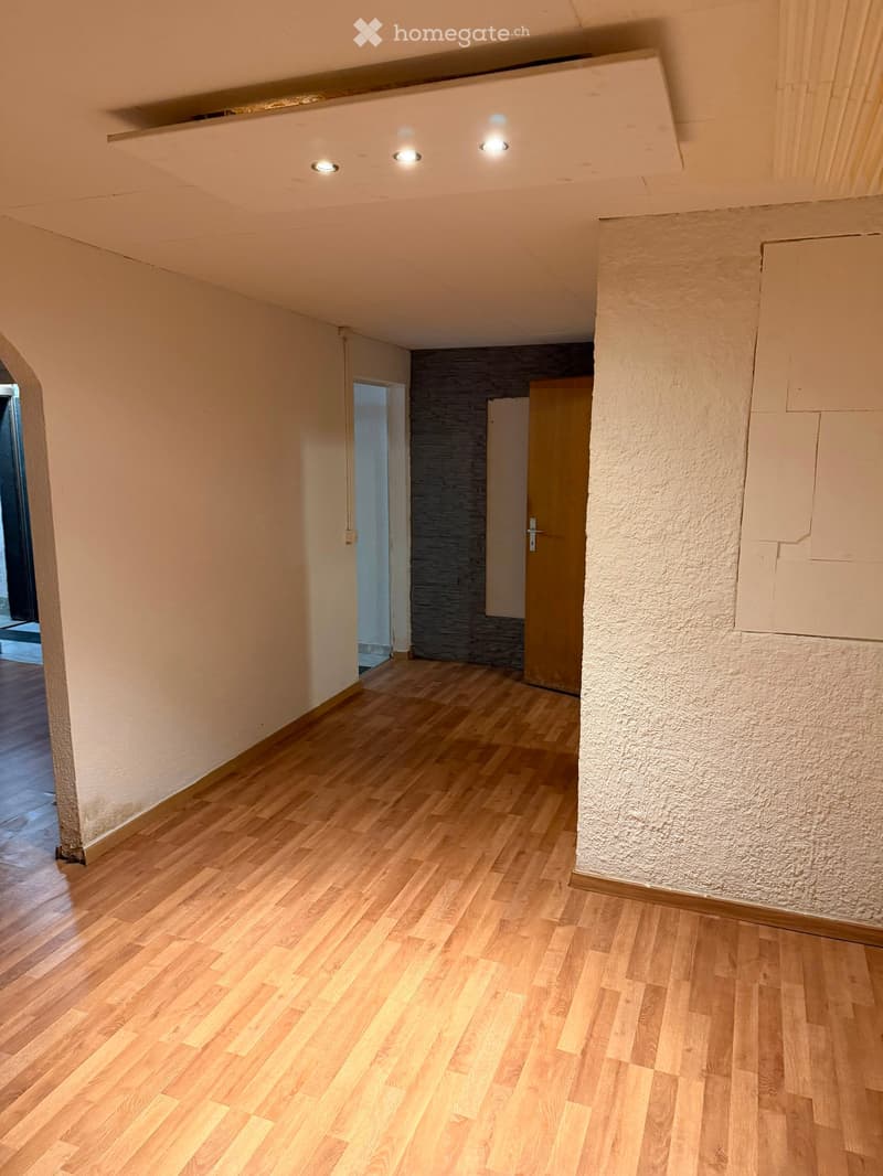 Empty hallway with wooden flooring, beige walls, and recessed ceiling lights. Features an arched doorway and a textured wall section.