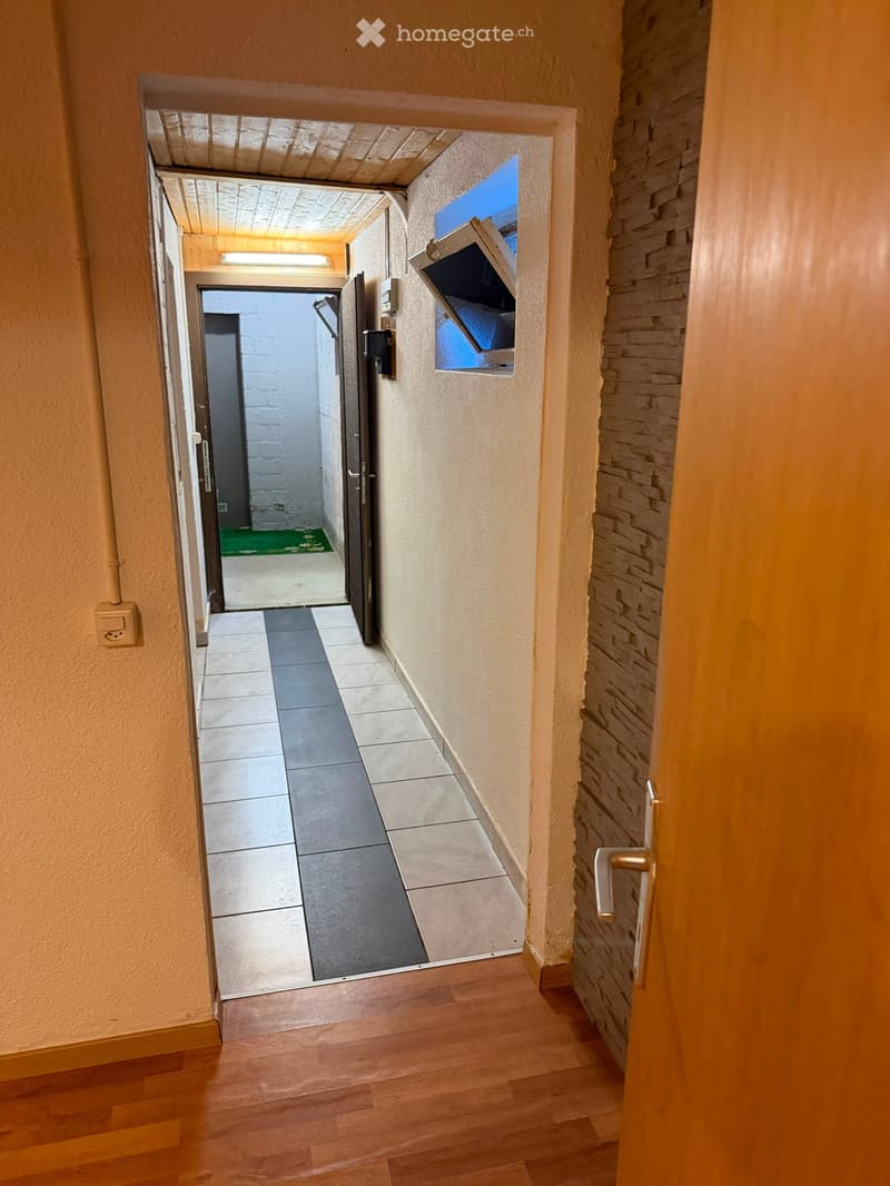 A narrow hallway with wooden flooring, beige walls, and a tiled section leading to another area. A door on the right and a partially open door at the end lead to other rooms.