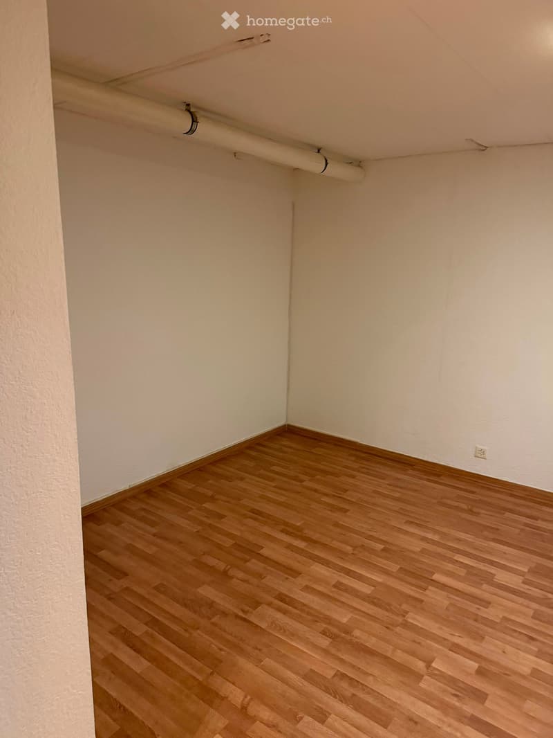 Empty room with light wooden flooring and white walls, featuring a visible electrical outlet and a small pipe running along the ceiling.