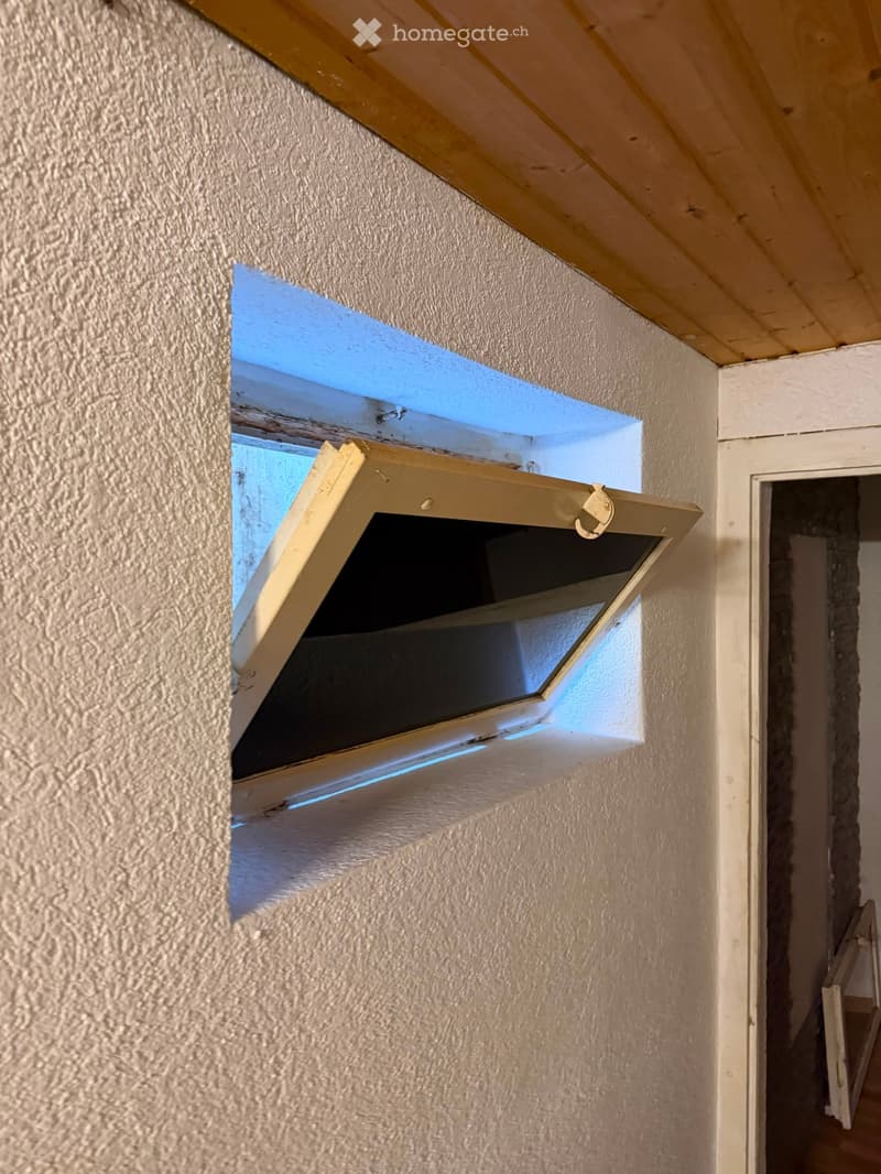 An unfurnished attic space with exposed wooden beams, a small window, and unfinished walls.