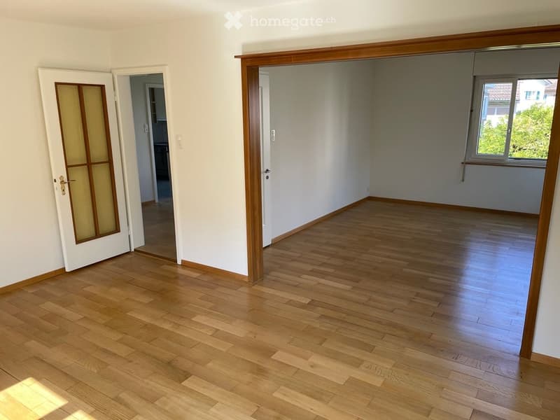 Empty room with wooden flooring, white walls, and a large window with a view of greenery. The room has a door with a glass panel leading to another area.