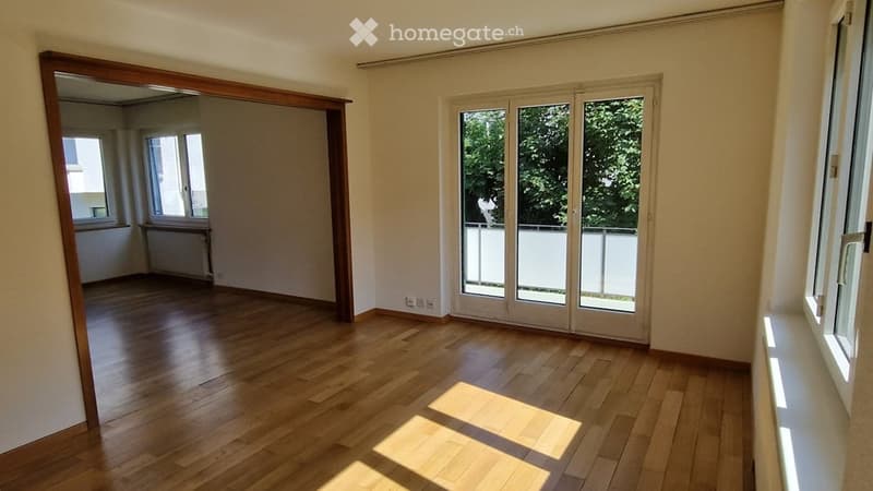 Empty room with wooden flooring, large windows, and a sliding glass door leading to a balcony area.