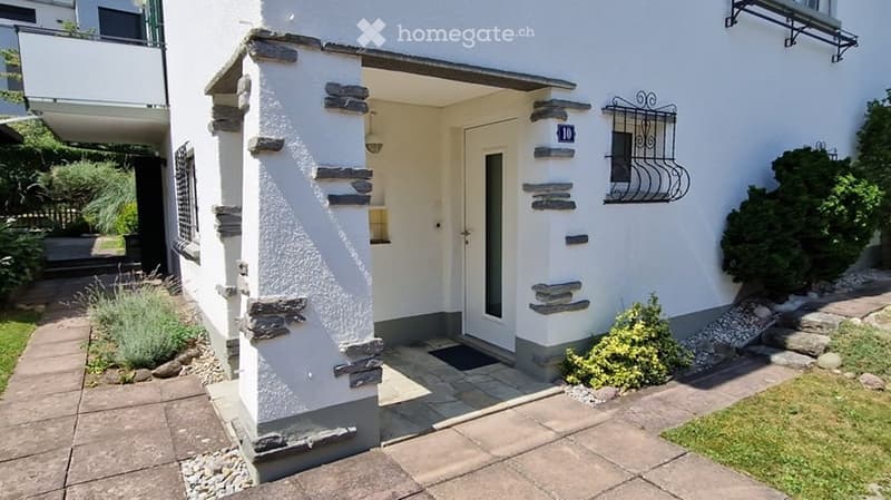 White building entrance with stone accents, small garden area, and decorative wrought iron window detail.