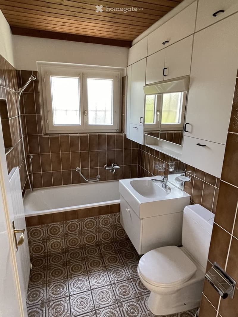 Bathroom with a bathtub, sink, toilet, mirror, and cabinet. Brown tiles on walls and floor, wooden ceiling, and a window providing natural light.