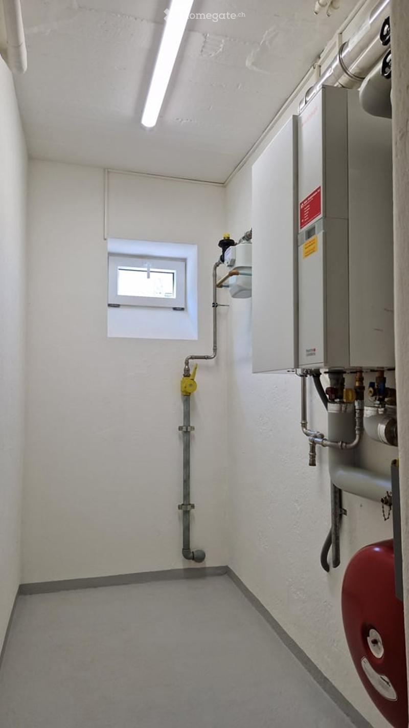 Utility room with white walls, grey floor, a small window, and various pipes and a water heater unit.