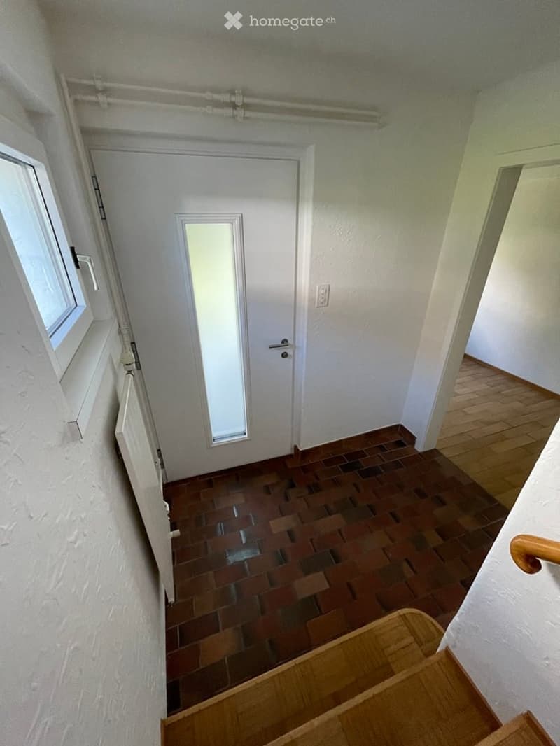Empty hallway with wooden flooring, a white door with a small window, and a staircase leading upwards.