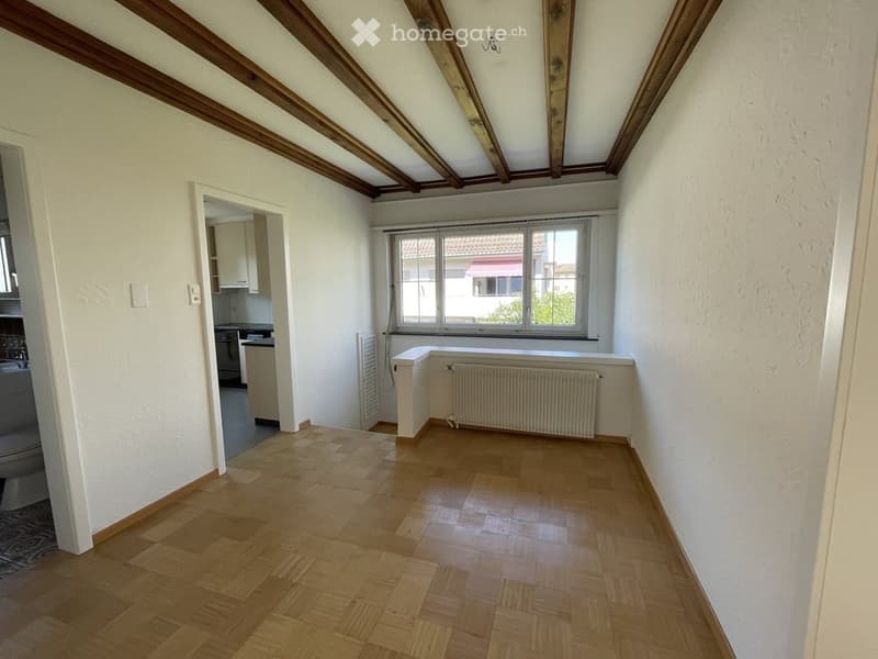 Empty room with wooden flooring, white walls, exposed wooden beams on the ceiling, and a window with a radiator below it. The room is unfurnished and has a simple, clean design.