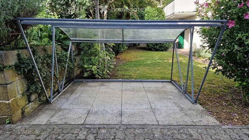 A covered outdoor patio with a metal frame and glass panels, surrounded by greenery and a stone wall. The floor is tiled, and there is a grassy area beyond the patio.