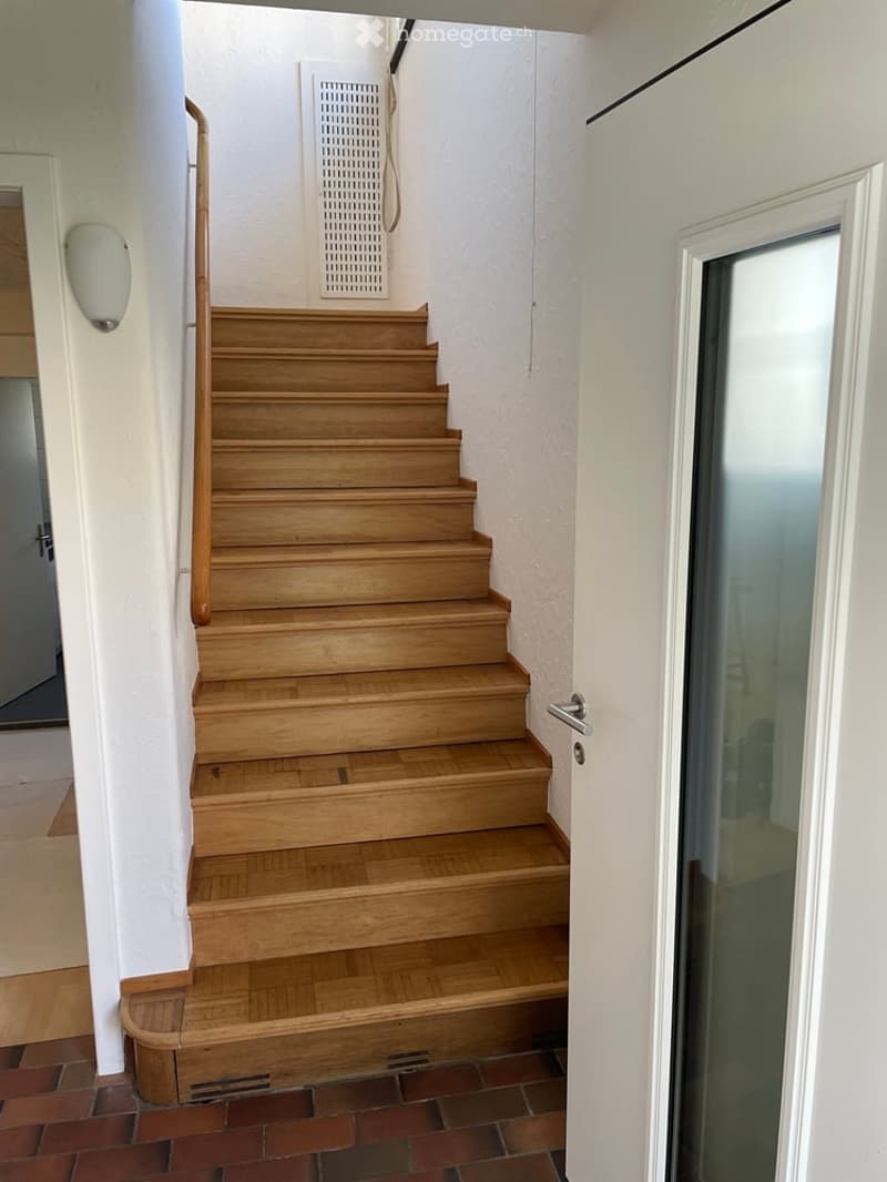 Wooden staircase with a handrail, white walls, and a light fixture on the wall. The floor is tiled with a mix of red and beige tiles.