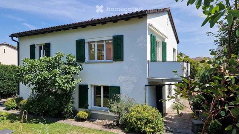 Two-story white house with green shutters, balcony, and well-maintained garden.