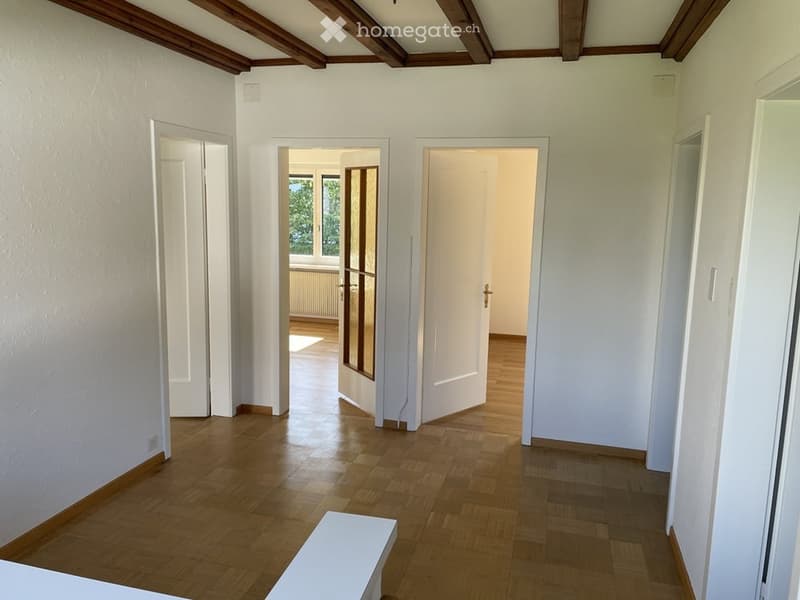 Empty hallway with wooden flooring, white walls, and several doors leading to other rooms. The ceiling features exposed wooden beams.