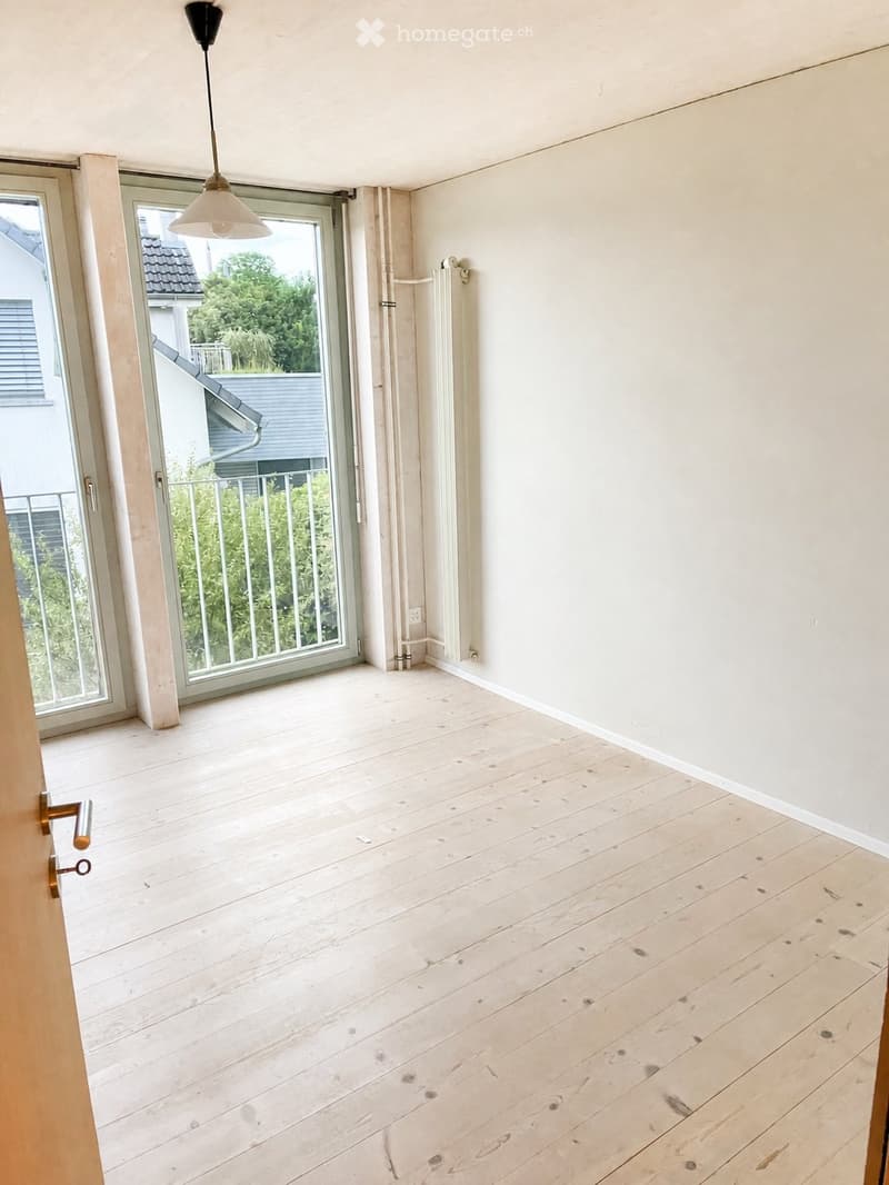 Empty room with light wooden flooring, white walls, large window with a view of greenery, and a hanging light fixture.