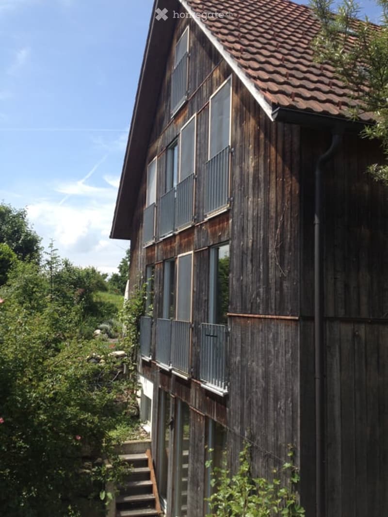 A two-story wooden house with large windows and a tiled roof, surrounded by greenery and trees.
