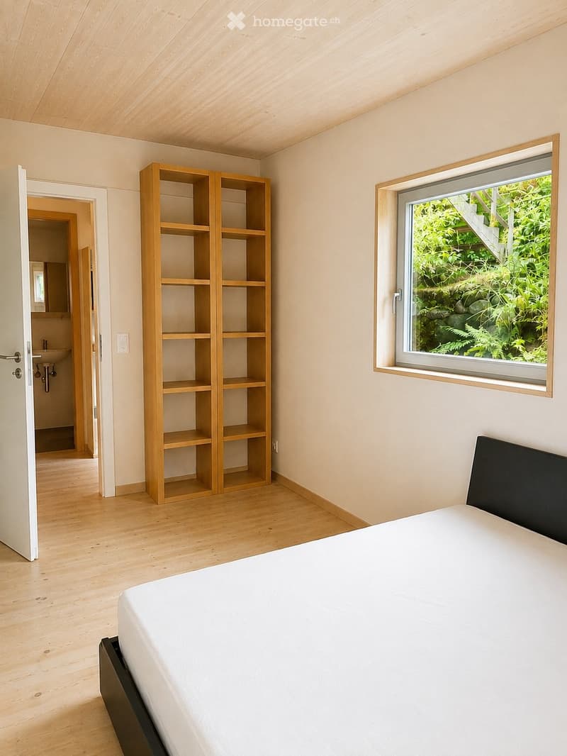 Bright bedroom with wooden flooring, a white bed with a black headboard, a wooden shelf unit, and a large window overlooking greenery.