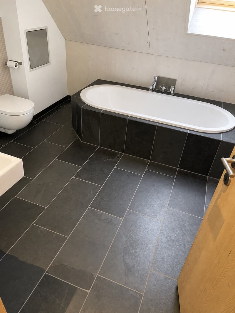 Modern bathroom with dark grey tiles, a white bathtub, a toilet, and a mirror on the wall.