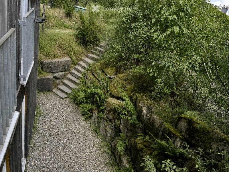 A raised garden path with stone steps leading up to a wooden deck, surrounded by lush greenery and ferns.