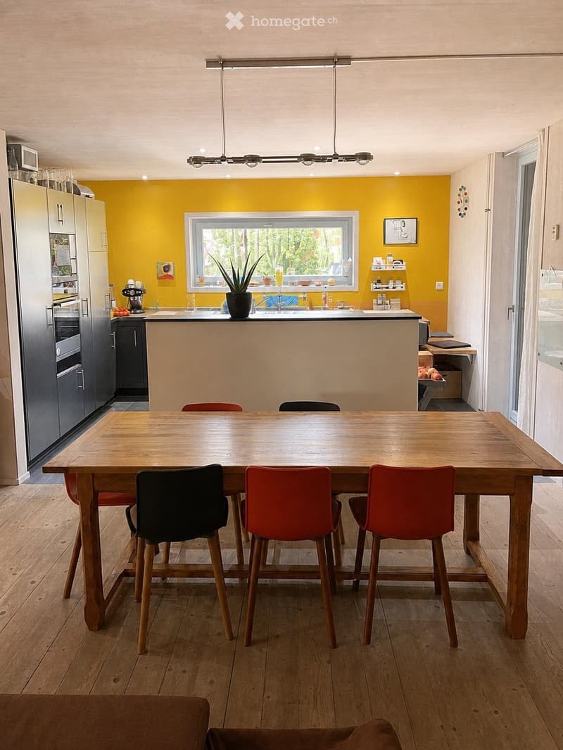 Bright kitchen with a yellow accent wall, wooden dining table, and modern appliances. Features include a bar counter, stools, and a potted plant on the counter.