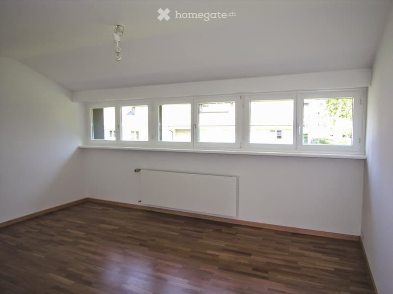 Empty room with wooden flooring, white walls, and large windows with white frames. A radiator is mounted on one wall.