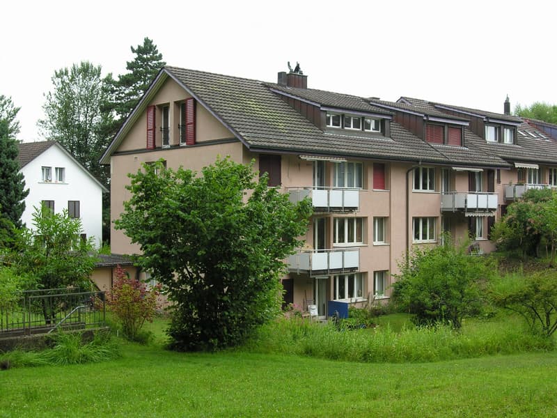 A two-story residential building with beige walls, red shutters, multiple windows, and balconies. The building is surrounded by a well-maintained lawn and trees.