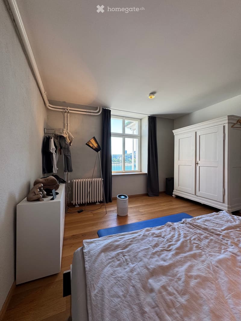 Bedroom with wooden floor, large window with dark curtains, bed with light-colored bedding, white wardrobe, floor lamp, radiator, air purifier, small table with a statue and other items.