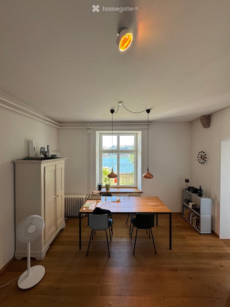 Bright room with wooden floor, large window offering a view, a table with chairs, a fan, a cabinet, a shelf with books, and a clock on the wall.