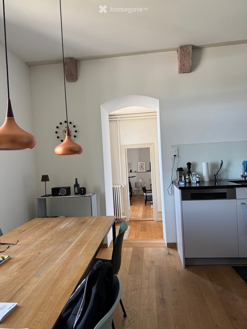 A modern, well-lit room with wooden flooring, a large wooden dining table, and a green chair. The room features a kitchen area with a sink, dishwasher, and various kitchen items. There is also a lamp, a radiator, and a small table with a lamp and a bottle