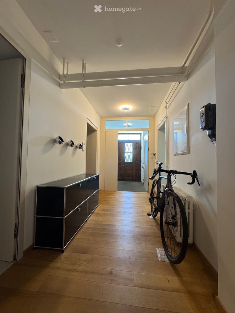 A modern hallway with wooden flooring, recessed lighting, and a bicycle leaning against the wall. A black cabinet is on the left, and a door leads to another room at the end.