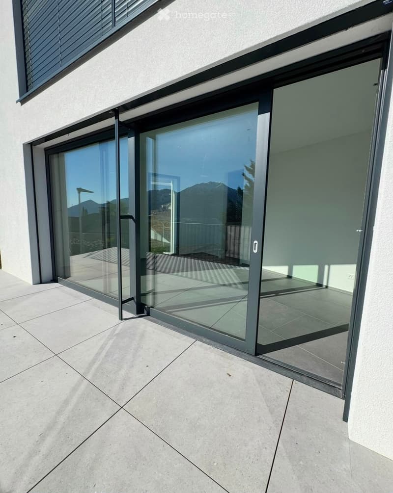 Modern glass sliding doors leading to a concrete tiled balcony, with a view of mountains in the background.