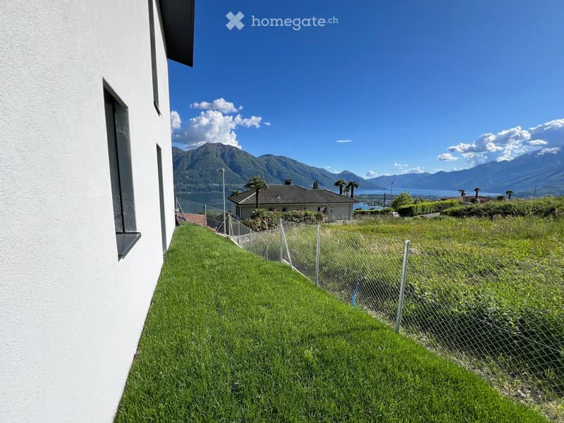 A grassy hillside with a metal fence, overlooking a scenic lake and mountain range in the distance.