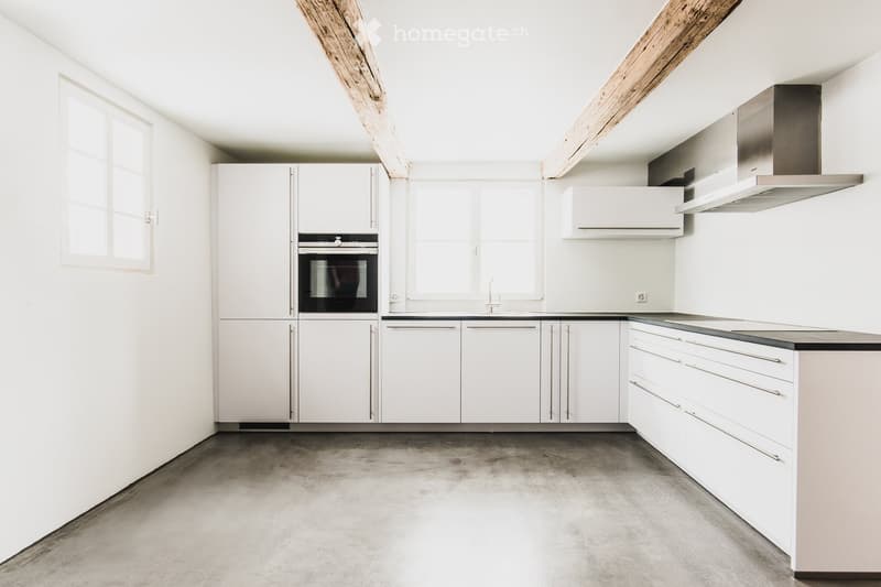 Modern, minimalist kitchen with white cabinets, stainless steel oven, range hood, and a concrete floor.