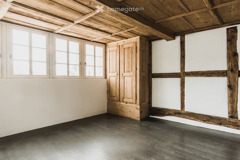 Empty room with exposed wooden beams, white walls, large windows, and a wooden cabinet.
