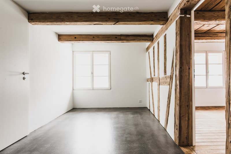 Empty room with white walls, exposed wooden beams, concrete floor, and large windows.
