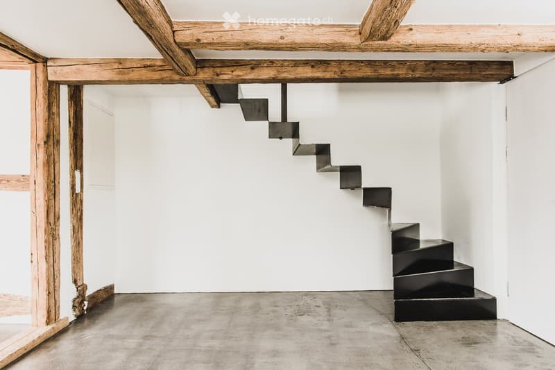 An empty hallway with exposed wooden beams, a modern black staircase, and a concrete floor.